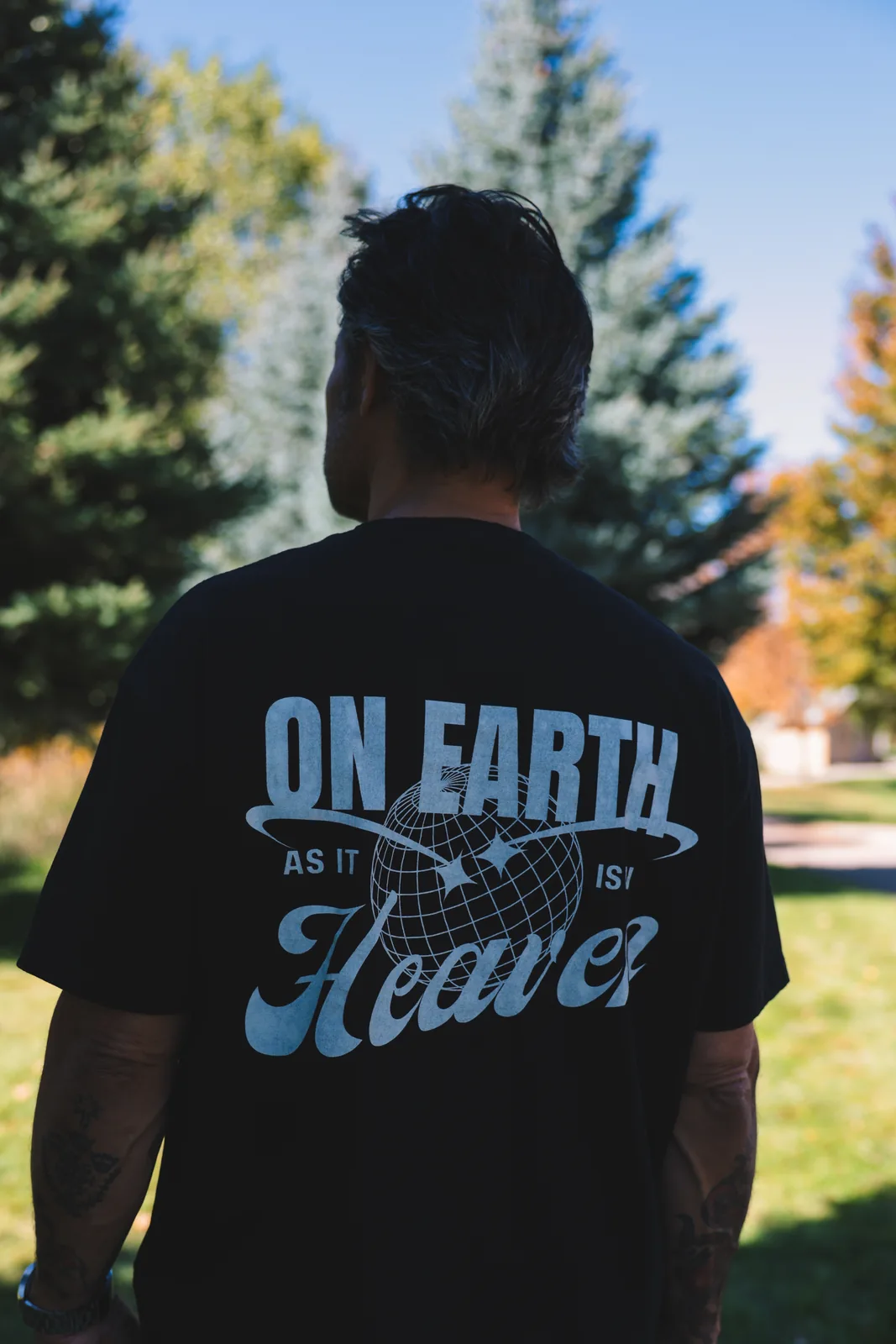 Back of a black tee with a globe graphic reading On Earth As It Is In Heaven