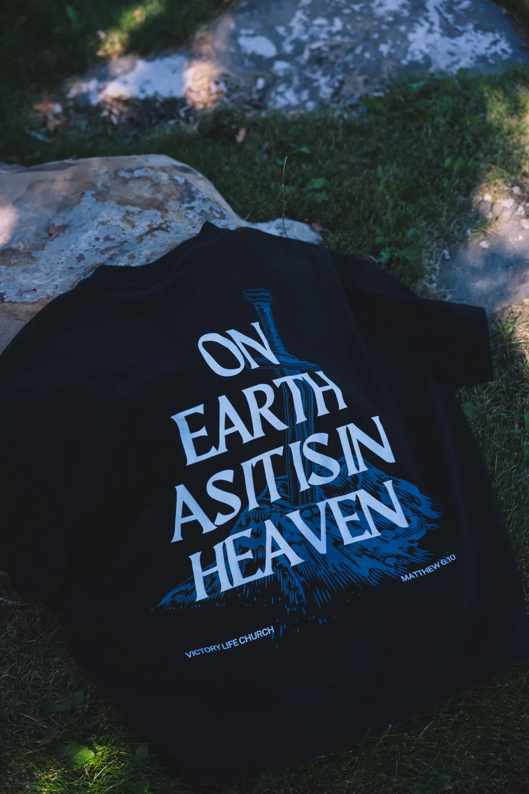 Black tee with a sword illustration reading On Earth As It Is In Heaven